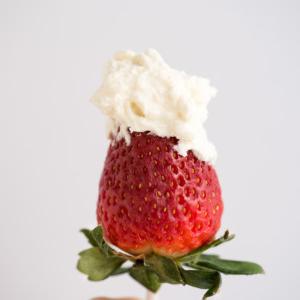 Una mano che tiene una fragola fresca su uno stecchino, guarnita con una generosa cucchiaiata di panna montata al cheesecake su uno sfondo chiaro e semplice.