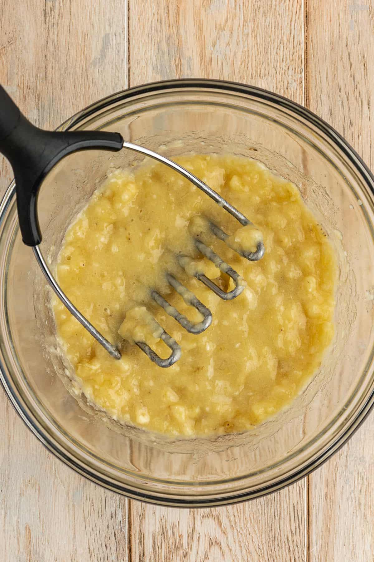 Una ciotola di vetro piena di banane schiacciate con uno schiacciapatate appoggiato nella ciotola, su una superficie di legno