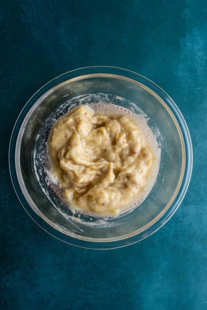 Banane schiacciate in una ciotola di vetro trasparente su una superficie color ottanio scuro.