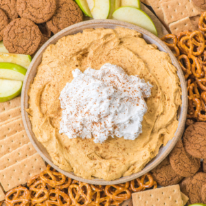 Una ciotola di cremosa crema di zucca con panna montata e cannella, circondata da fette di mela verde, pretzel, cracker Graham e biscotti allo zenzero.