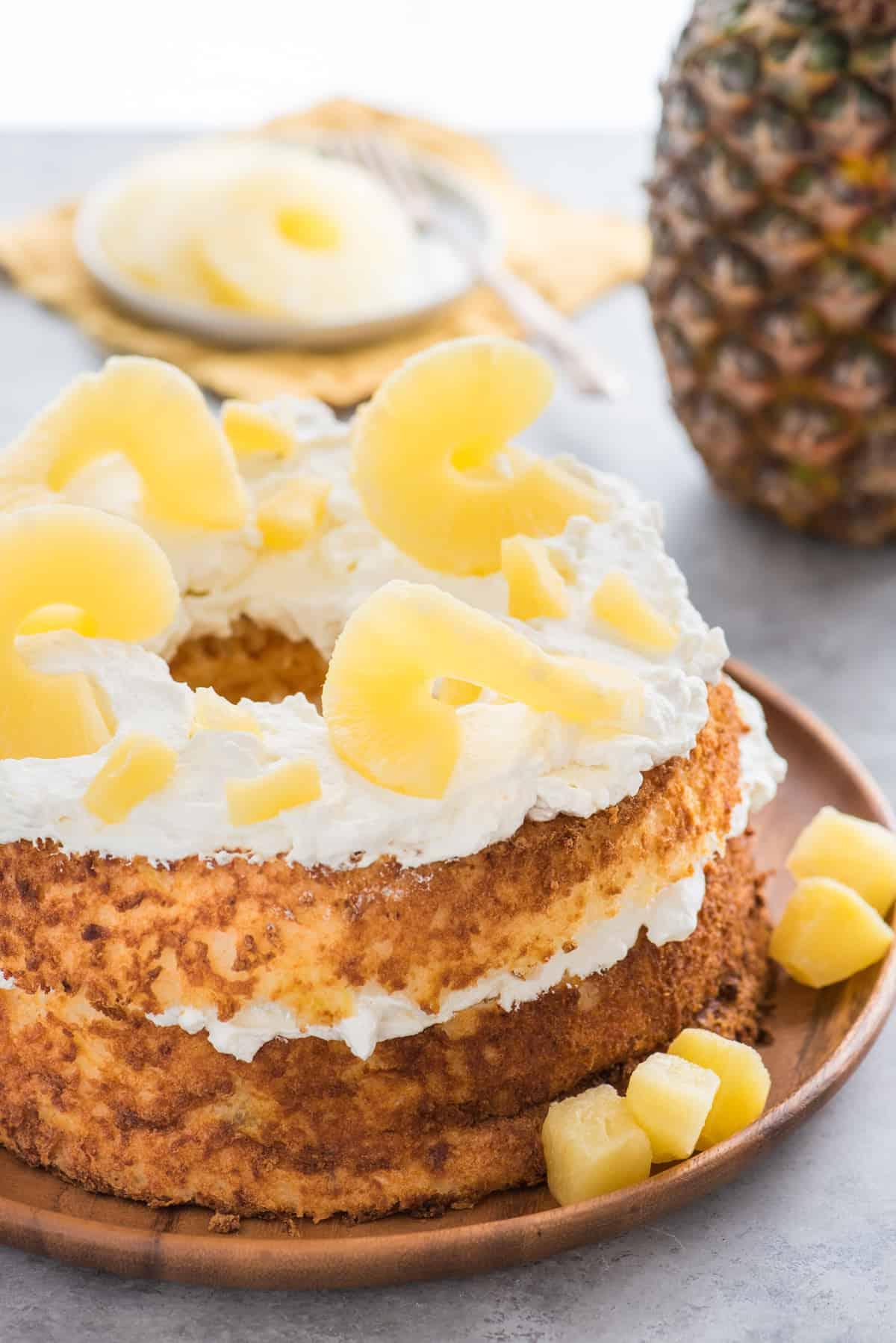 Torta degli angeli all'ananas 8 Torta Angel Food intera guarnita con panna montata e fette di ananas su vassoio di legno su sfondo bianco
