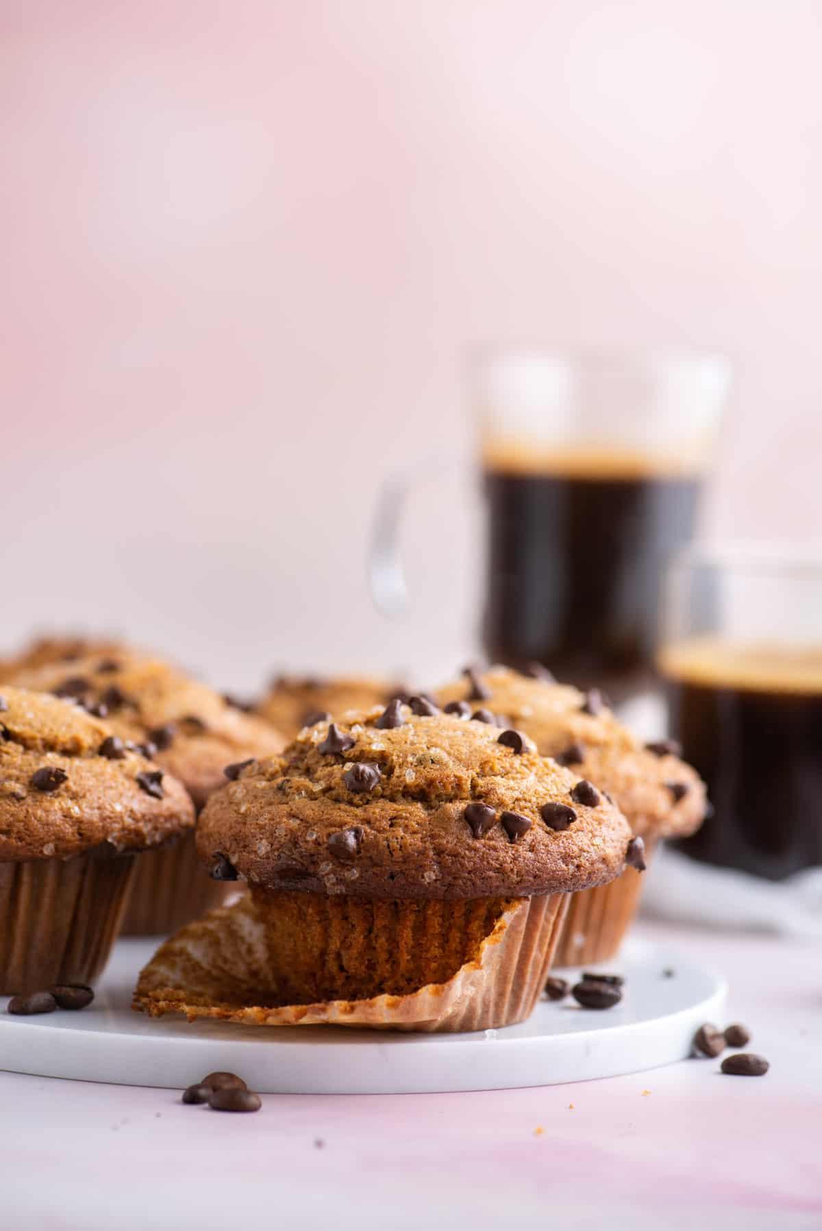 Muffin al Cappuccino e Gocce 8 muffin al cappuccino con gocce di cioccolato su un piatto bianco, appoggiati nel loro pirottino aperto, con chicchi di caffè sparsi intorno, altri muffin e due tazze di caffè sullo sfondo