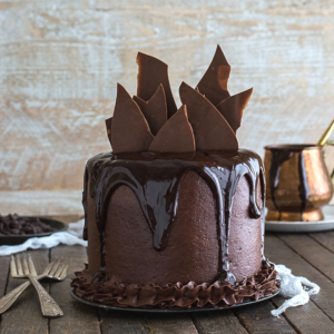 Una torta alta al cioccolato con glassa lucida che cola dai lati, guarnita con grandi scaglie di cioccolato, su un tavolo di legno con forchette e una pentola di rame sullo sfondo.