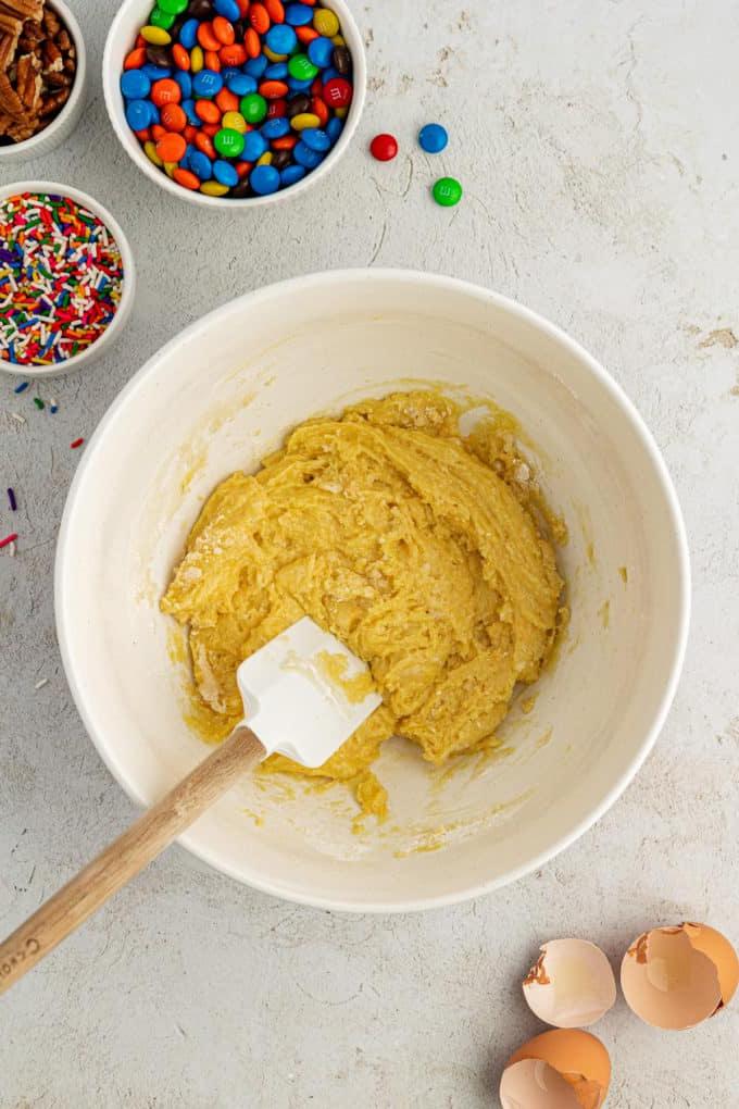 Impasto per biscotti con preparato per torte in una ciotola bianca con una spatola bianca e di legno circondata da gusci d'uovo, una ciotola di codette, una ciotola di M&M's e una ciotola di noci
