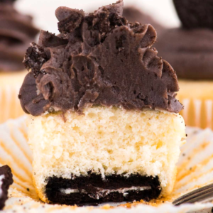 Un cupcake con fondo Oreo, con impasto alla vaniglia, un biscotto al cioccolato alla base e glassa al cioccolato spessa in cima. Il cupcake è in un pirottino di carta bianco e ne è stato morso un pezzo, rivelando i suoi deliziosi strati.