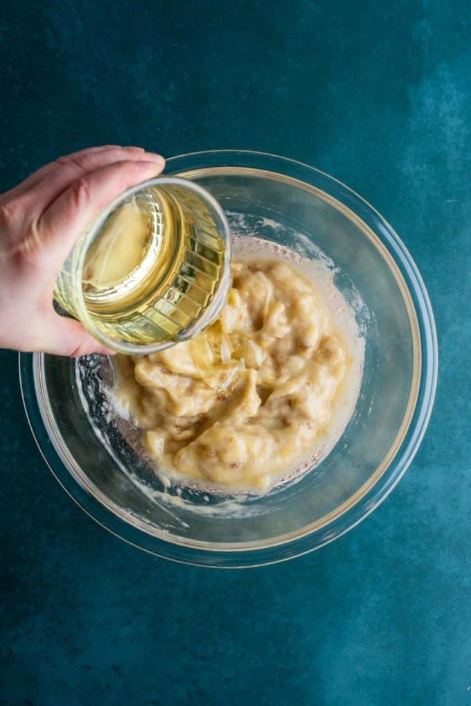 Banane schiacciate in una ciotola di vetro trasparente su una superficie color ottanio scuro con un piatto di vetro di olio vegetale che viene versato.
