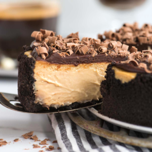 Un primo piano di una cremosa cheesecake al caffè con crosta al cioccolato e scaglie di cioccolato in cima. Una fetta viene sollevata, rivelando strati lisci e una ricca consistenza. La torta decadente poggia su un panno a strisce.