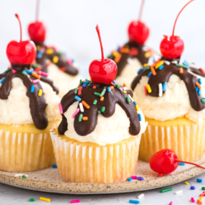 Vanilla ice cream cupcakes topped with white frosting, chocolate drizzle, rainbow sprinkles, and a red cherry on top, arranged on a plate with extra cherries and sprinkles scattered around.