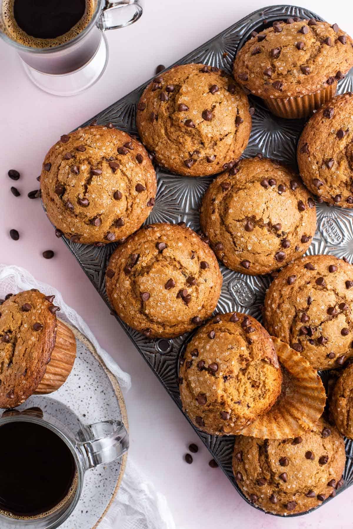 Muffin al Cappuccino e Gocce 9 vista dall'alto di una teglia per muffin piena di muffin al cappuccino con gocce di cioccolato con chicchi di caffè sparsi intorno, con una tazza di caffè e un piatto con un muffin e una tazza di caffè accanto