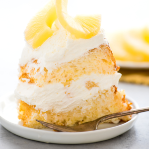 Torta degli angeli all'ananas 9 Una fetta di soffice torta Angel Food all'ananas a strati con panna montata e guarnita con fette di ananas su un piatto bianco con una forchetta accanto.
