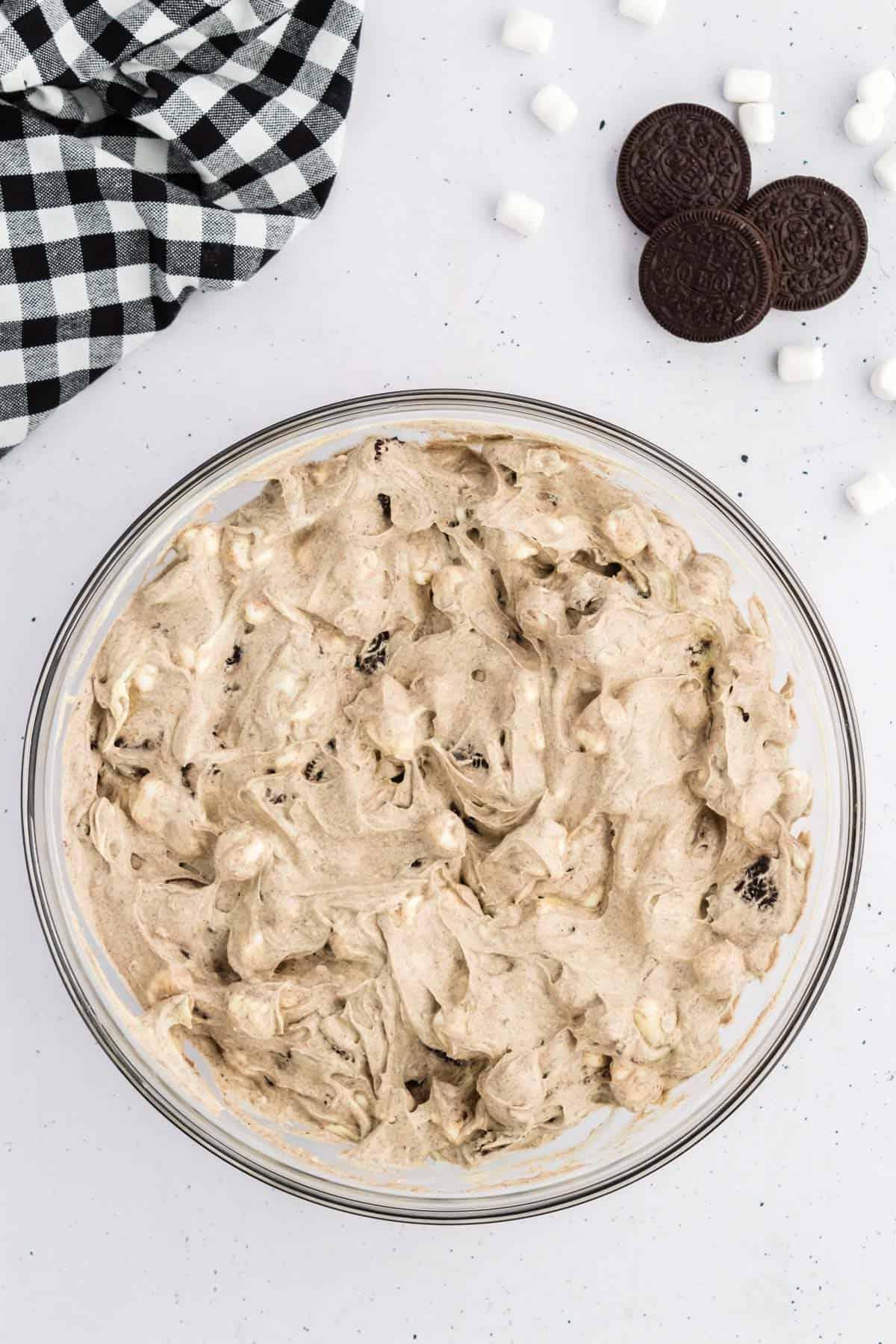 Fluff di Oreo in una ciotola di vetro circondato da Oreo, mini marshmallow e un panno da cucina a scacchi bianchi e neri