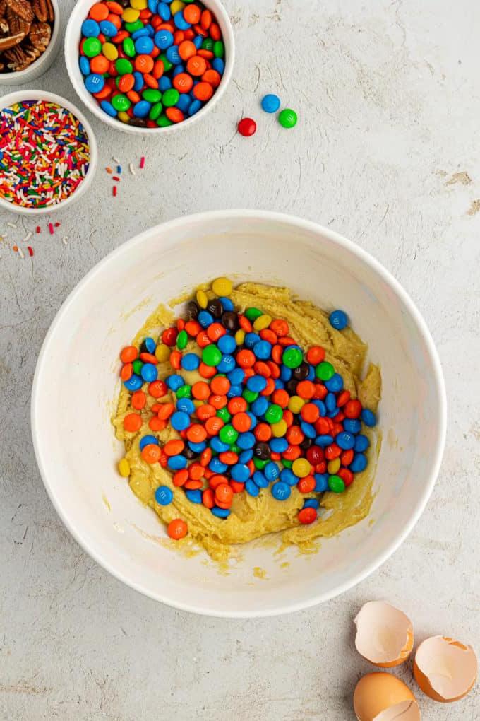 Impasto per biscotti con preparato per torte in una ciotola bianca con un mucchio di M&M's sopra, circondato da gusci d'uovo, una ciotola di codette, una ciotola di M&M's e una ciotola di noci