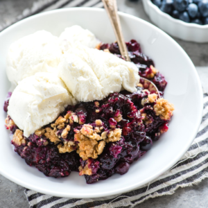 Una ciotola bianca di Crumble ai Mirtilli sormontata da gelato alla vaniglia, appoggiata su un panno a righe, accompagnata da un cucchiaio e una ciotola di mirtilli freschi sullo sfondo.