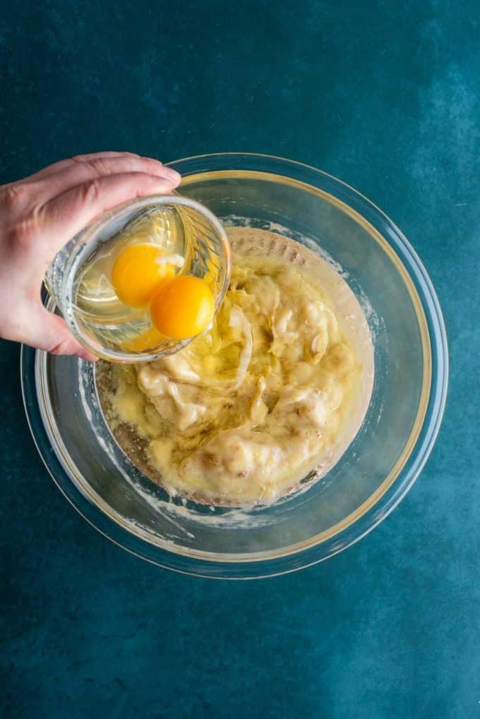 Banane schiacciate e olio vegetale in una ciotola di vetro trasparente su una superficie color ottanio scuro con due uova che vengono versate sopra.