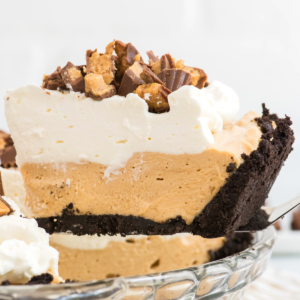 Una fetta di cremosa torta fredda al burro di arachidi senza cottura con una crosta di biscotti al cioccolato, guarnita con panna montata e pezzi di burro di arachidi tritati, sollevata da una teglia di vetro.