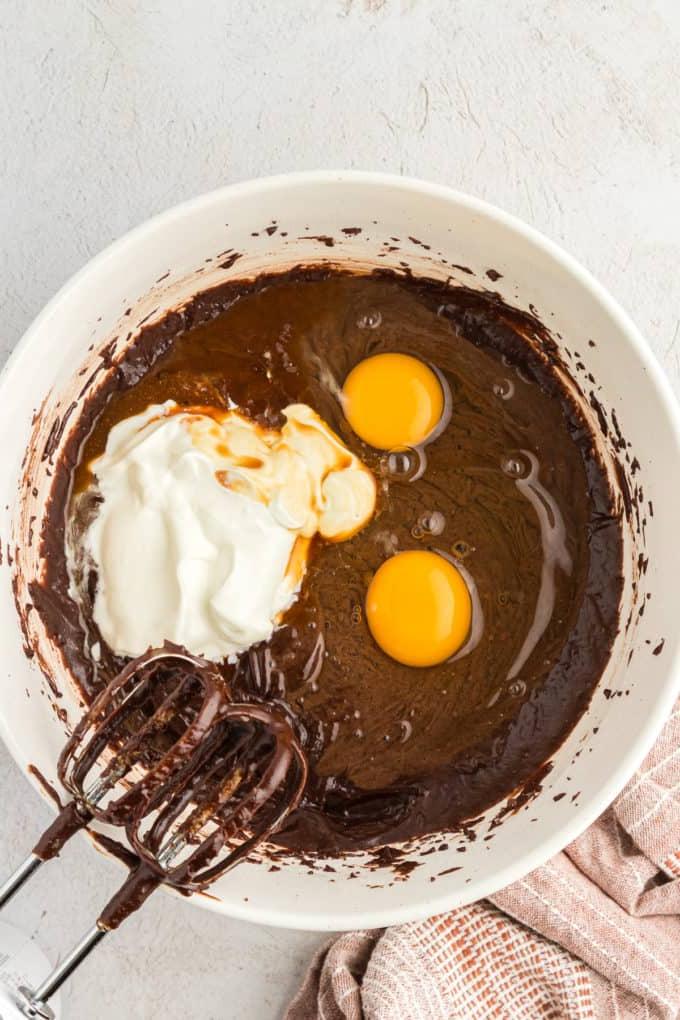 Impasto per torta Texana in una ciotola bianca con due uova crude e panna acida sopra, mixer coperti di impasto al cioccolato appoggiati alla ciotola e un panno da cucina color cuoio a sinistra della ciotola