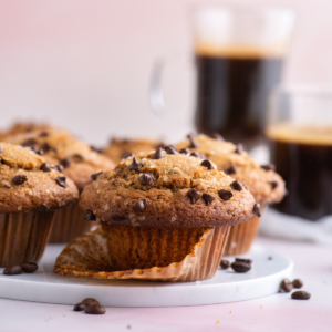 Muffin al Cappuccino e Gocce 10 muffin al cappuccino con gocce di cioccolato su un piatto bianco con gocce di cioccolato sparse intorno e due tazze di cappuccino sullo sfondo