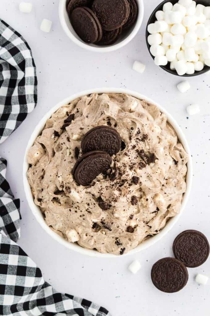 Fluff di Oreo sormontato da briciole di Oreo e due Oreo interi in una ciotola di vetro circondato da Oreo, mini marshmallow e un panno da cucina a scacchi bianchi e neri