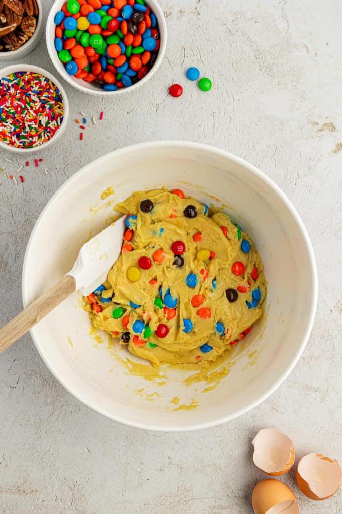 Impasto per biscotti con preparato per torte in una ciotola bianca con con M&M's mescolati con una spatola bianca e di legno circondata da una ciotola di codette, una ciotola di M&M's e una ciotola di noci