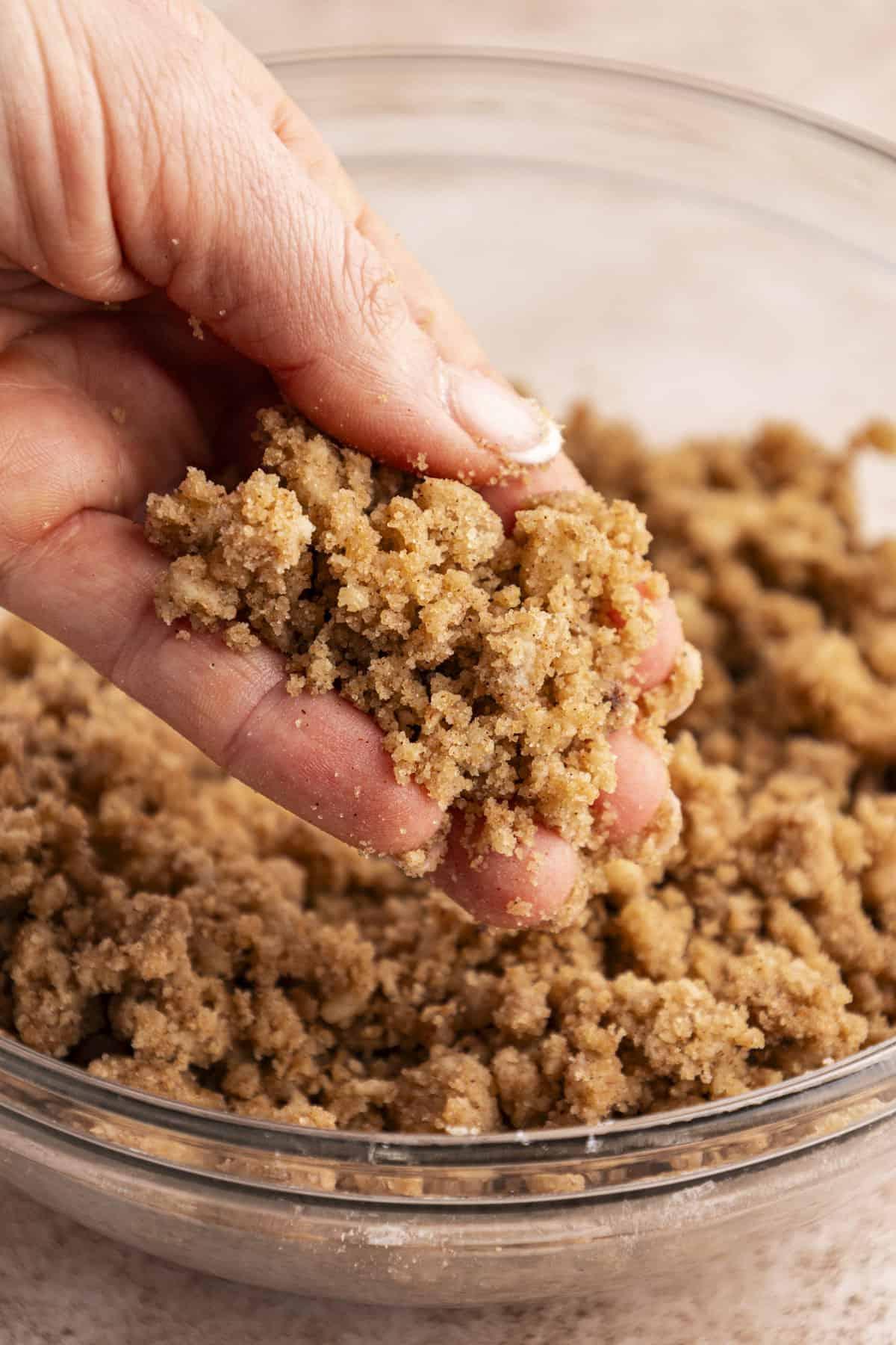 Una mano che tiene un topping croccante sopra una ciotola di vetro trasparente piena di altro topping croccante.