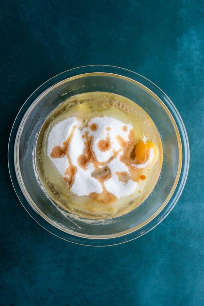 Ingredienti liquidi per il pane alle banane e gocce di cioccolato in una ciotola di vetro trasparente su una superficie color ottanio scuro, tra cui banane schiacciate, olio vegetale, uova, zucchero semolato ed estratto di vaniglia.
