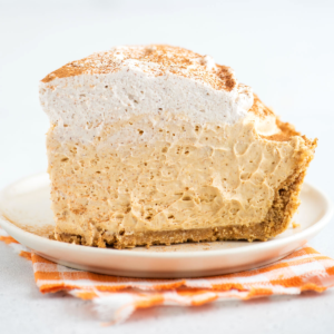 Un pezzo leggero e cremoso di torta di zucca senza cottura adagiato su un piatto da dessert, sopra un tovagliolo a quadretti arancioni.