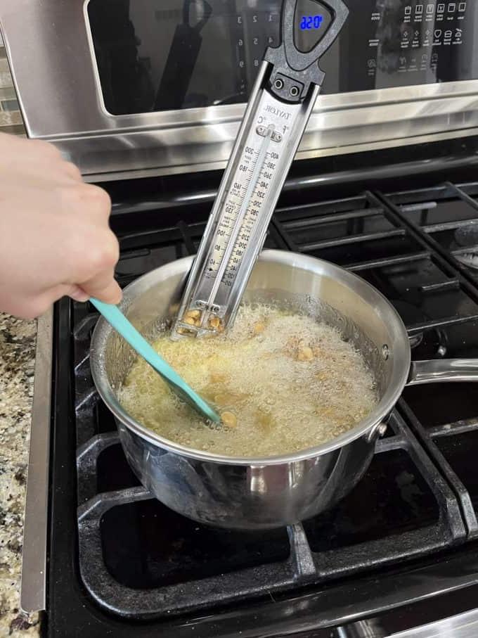 Una pentola sul fornello con zucchero semolato, acqua, sciroppo di mais e arachidi che cuociono, mescolate con una spatola di gomma e un termometro per dolci attaccato al lato