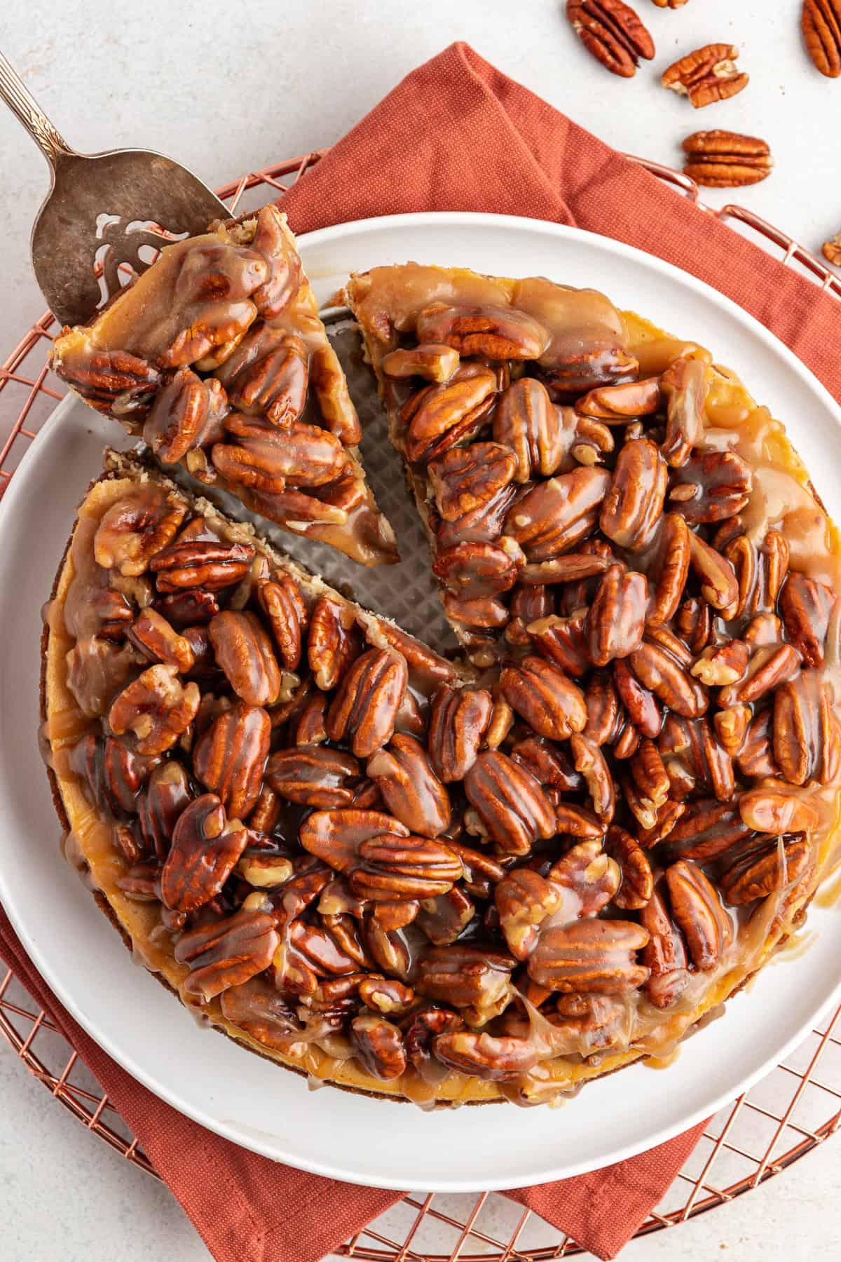Vista dall'alto di una cheesecake alle noci pecan su un panno arancione bruciato su una griglia, con una fetta rimossa da una spatola da portata e noci pecan sparse intorno