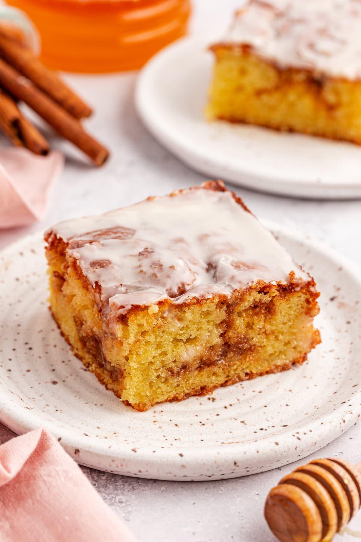 Una fetta di torta Honey Bun quadrata con glassa, poggiata su un piatto bianco maculato. La torta umida presenta spirali di cannella visibili. Sullo sfondo, parzialmente visibili, un'altra fetta, bastoncini di cannella e un mestolo per miele.