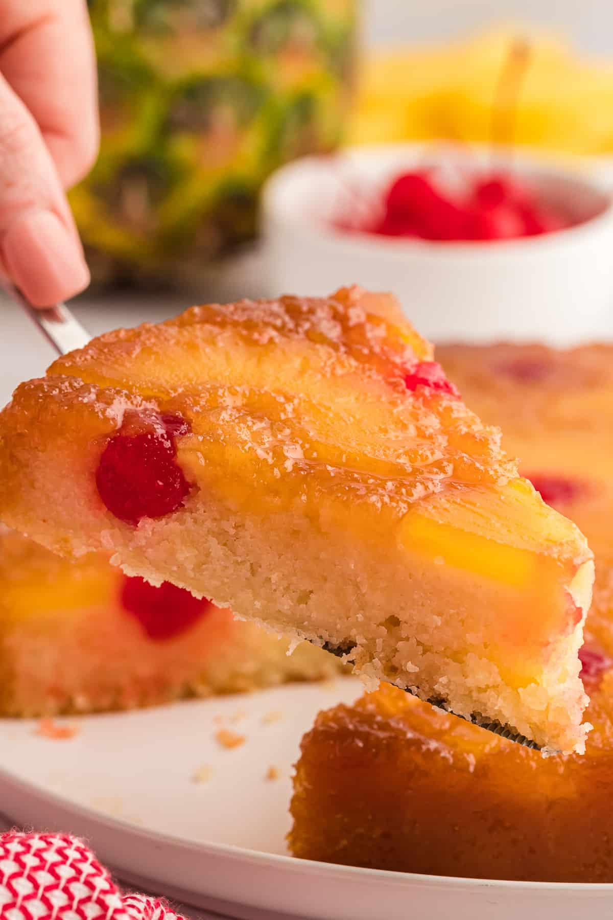 Una fetta di torta rovesciata all'ananas viene sollevata da un piatto, con un ananas intero, ciliegie e fette di ananas nello sfondo e un canovaccio rosso e bianco accanto al piatto.
