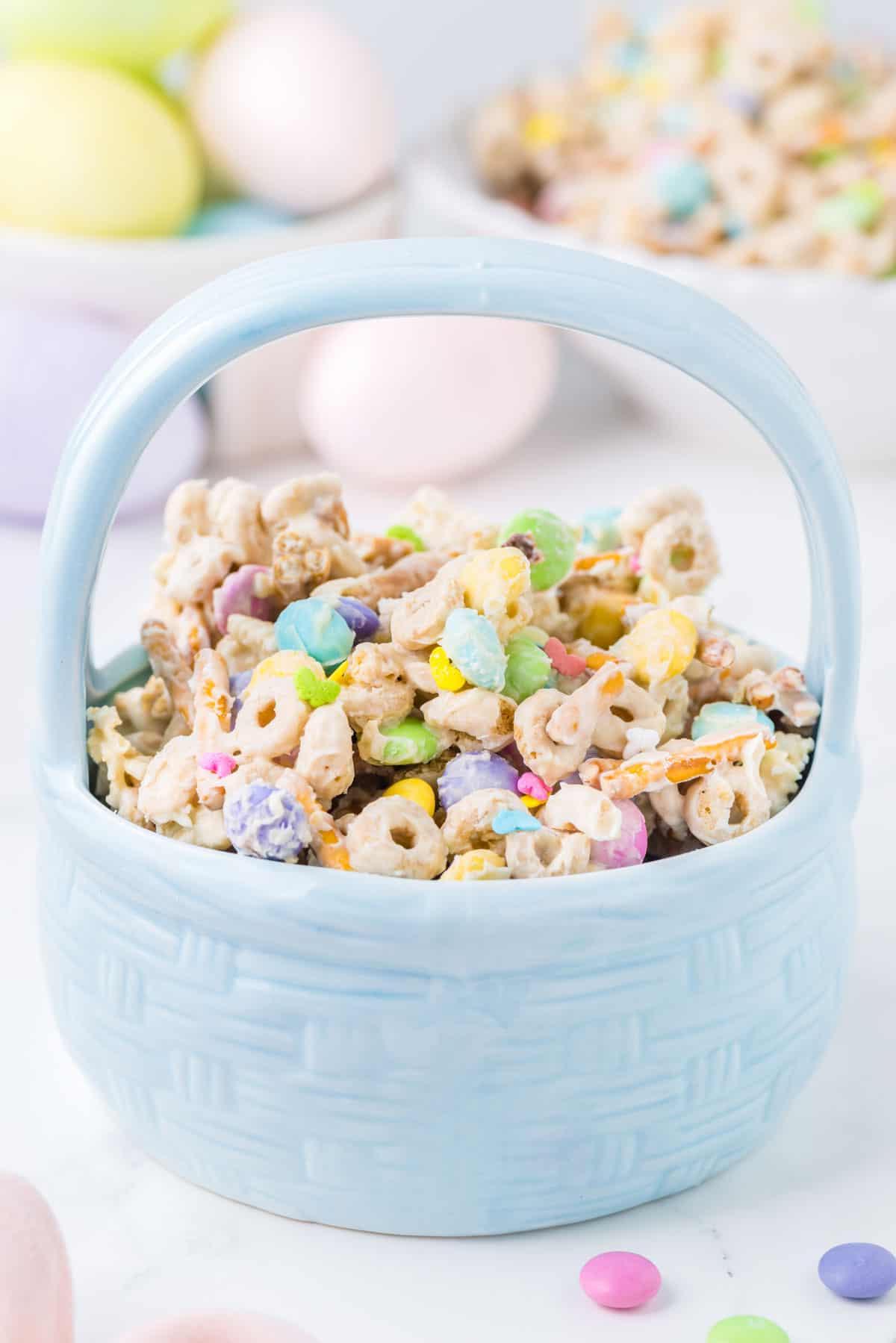 Snack mix 'Bunny Bait' pasquale in una ciotola blu a forma di cestino pasquale con uova di Pasqua e altro 'Bunny Bait' sullo sfondo e alcune M&M's intorno