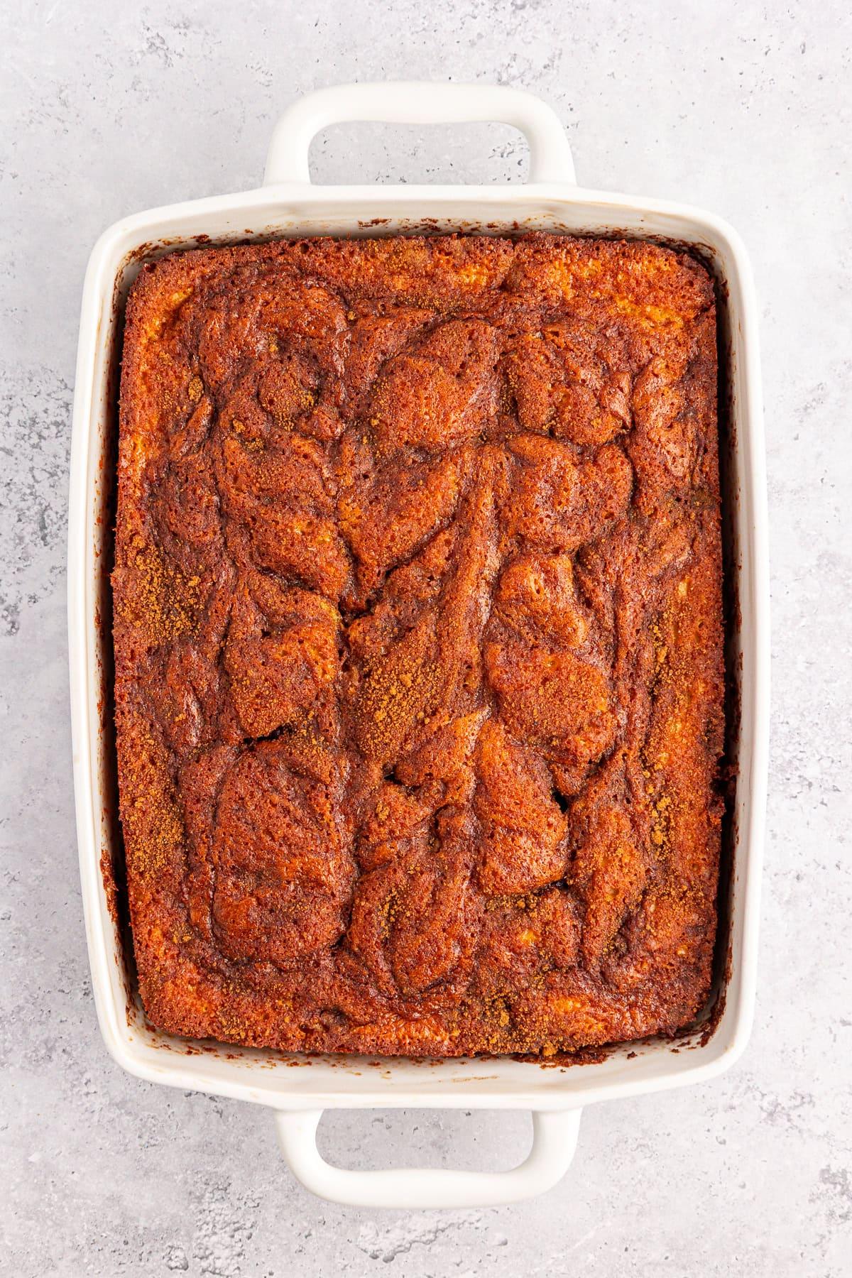 Una torta Honey Bun con una cima marmorizzata riposa in una teglia rettangolare bianca su uno sfondo grigio chiaro. La delizia di cannella cotta vanta una crosta dorata con spirali più scure, incarnando il perfetto connubio di dolcezza e calore.