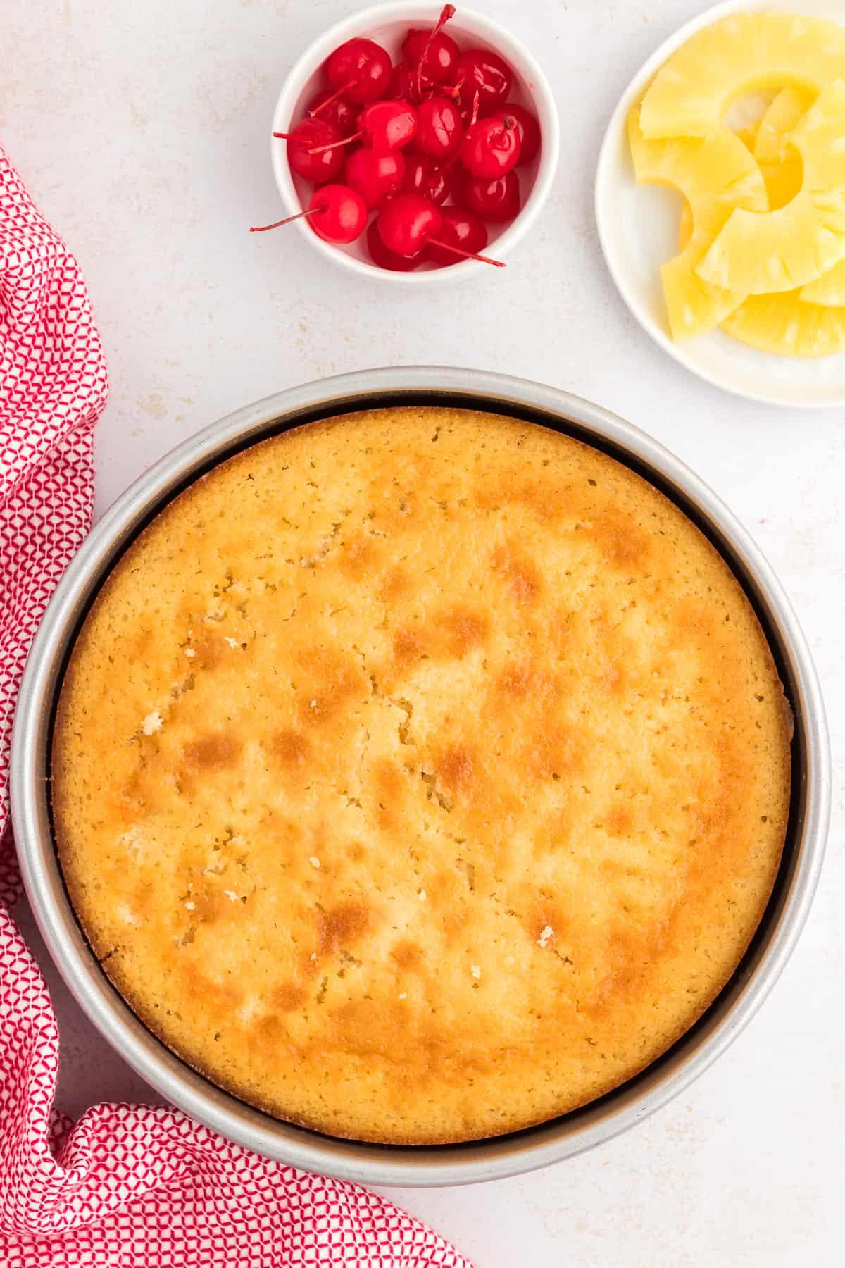 Torta rovesciata all'ananas appena sfornata, prima di essere capovolta, accanto a un piatto di fette di ananas e una ciotola di ciliegie al maraschino e un canovaccio rosso e bianco.