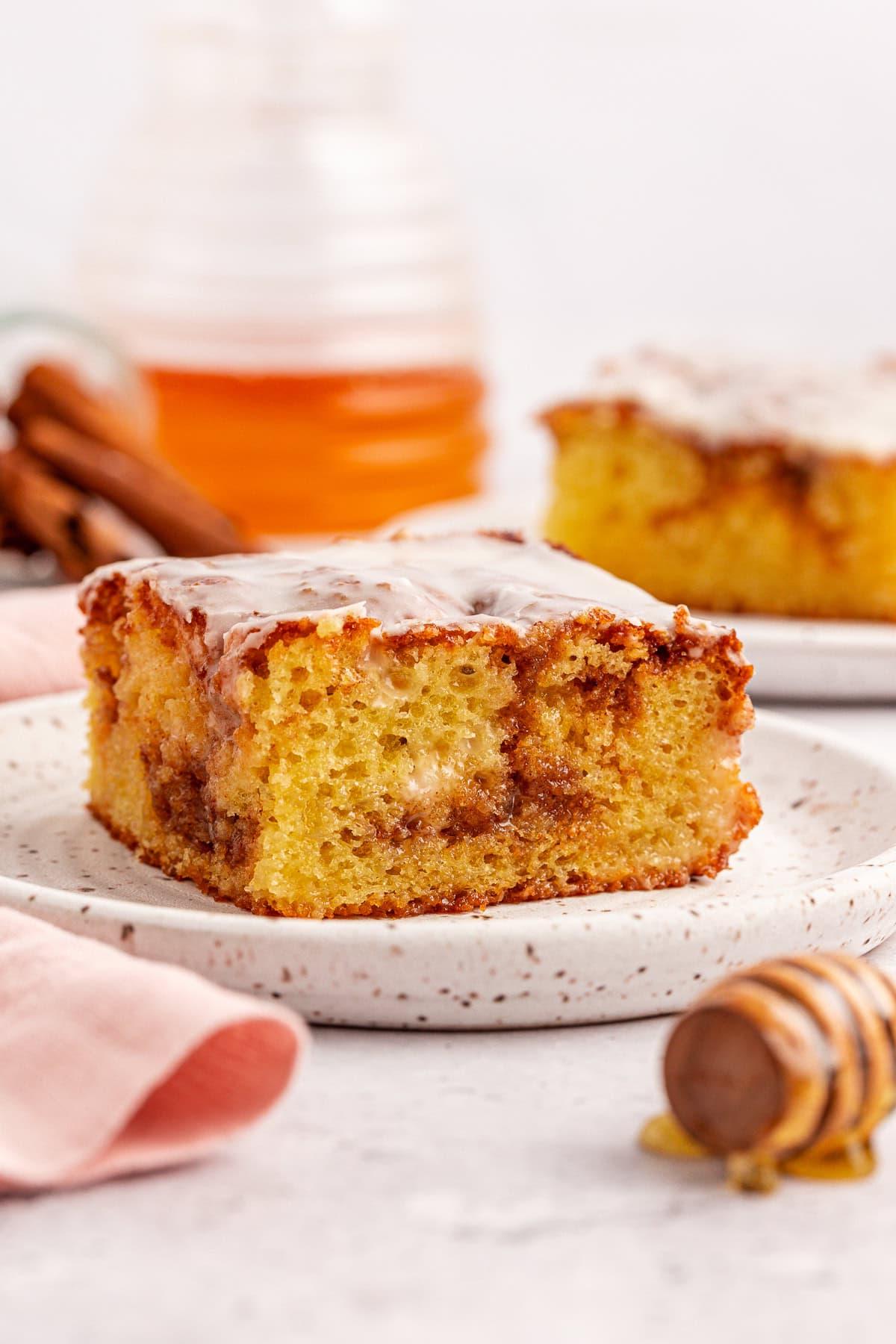 Primo piano di una fetta di torta di girelle alla cannella con glassa su un piatto maculato. Sullo sfondo, un'altra fetta di torta Honey Bun è su un piatto, e un mestolo per miele con gocce di miele è visibile. Un panno rosa chiaro è parzialmente mostrato.