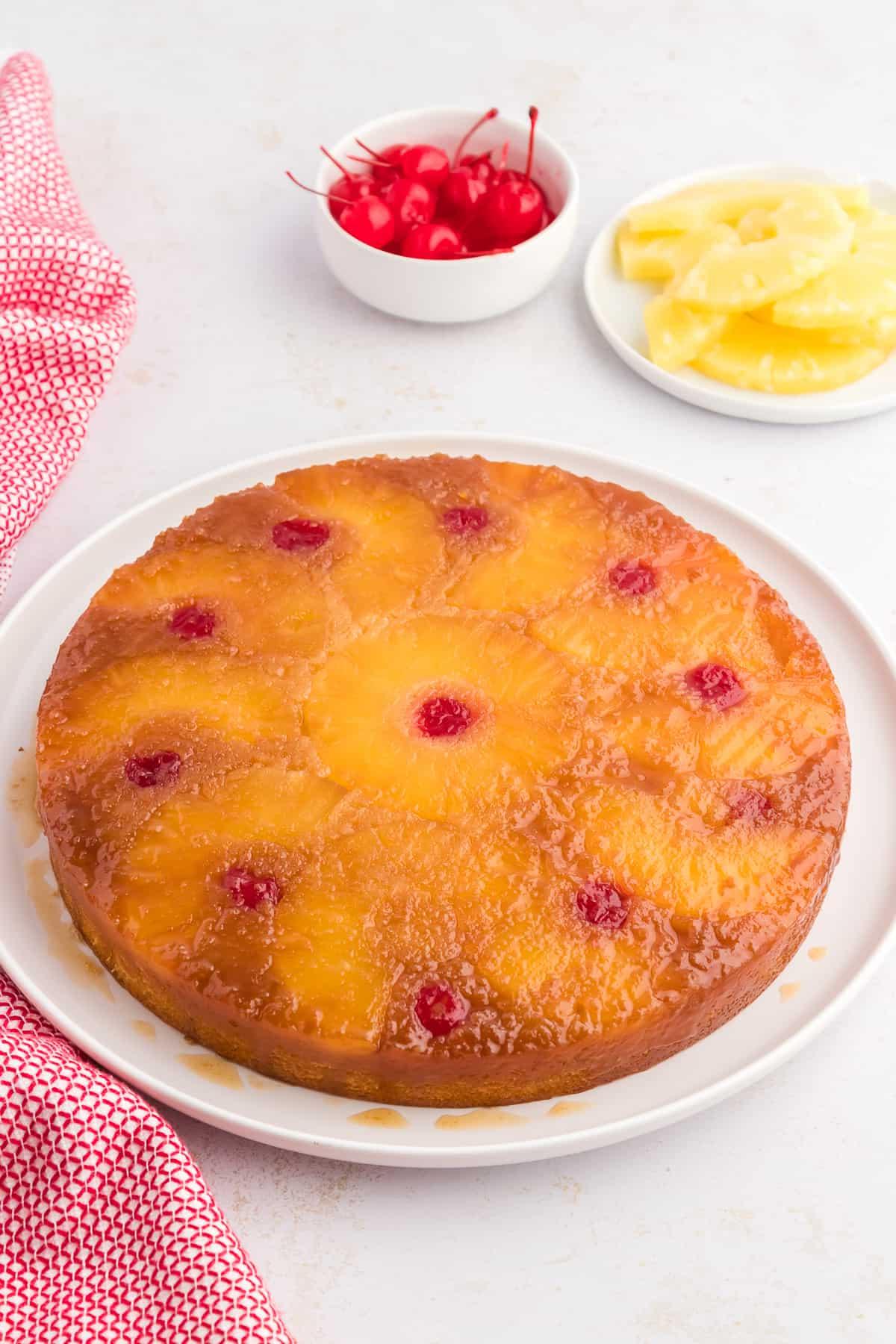 Torta rovesciata all'ananas su un piatto bianco circondata da fette di ananas, ciliegie al maraschino e un canovaccio rosso e bianco.