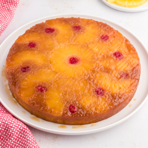 Torta rovesciata all'ananas su un piatto bianco circondata da fette di ananas, ciliegie al maraschino e un canovaccio rosso e bianco.