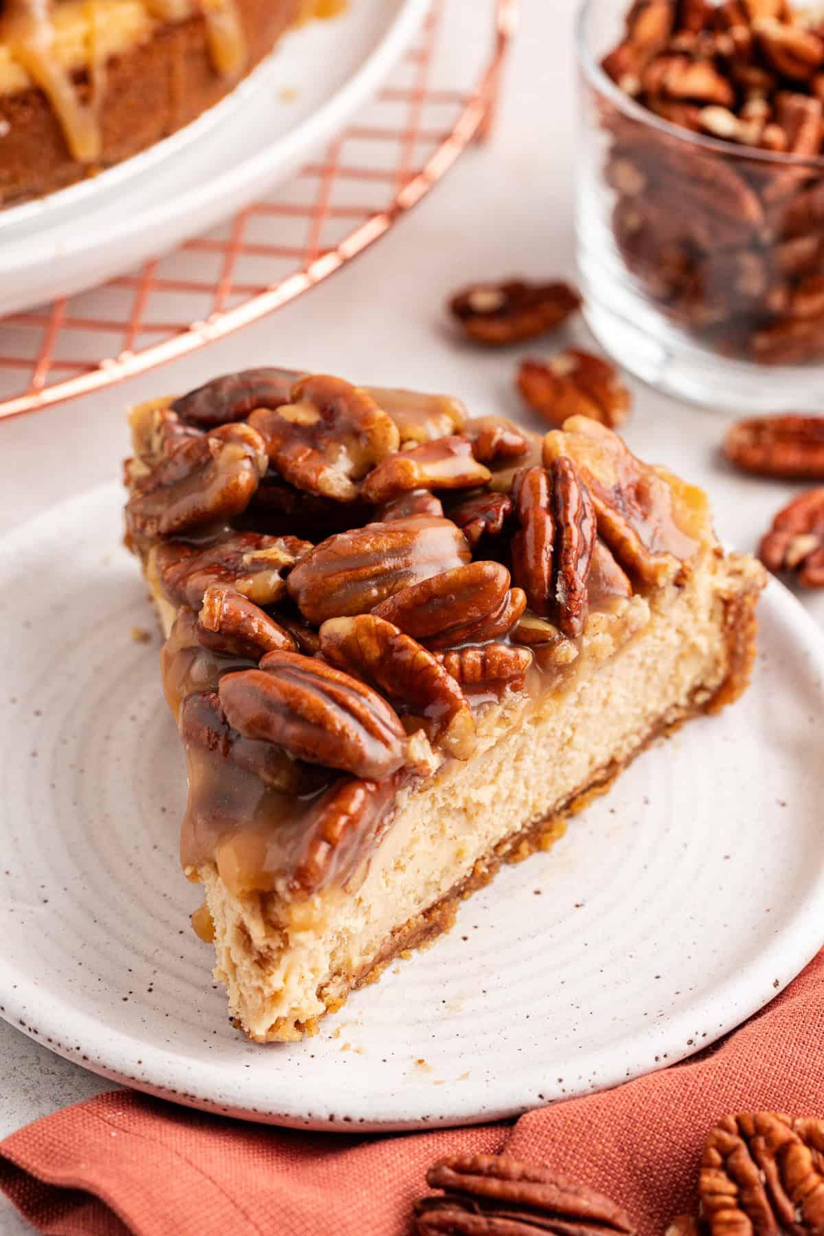 Una fetta di cheesecake alle noci pecan su un piattino bianco accanto a un panno arancione bruciato con noci pecan sparse intorno