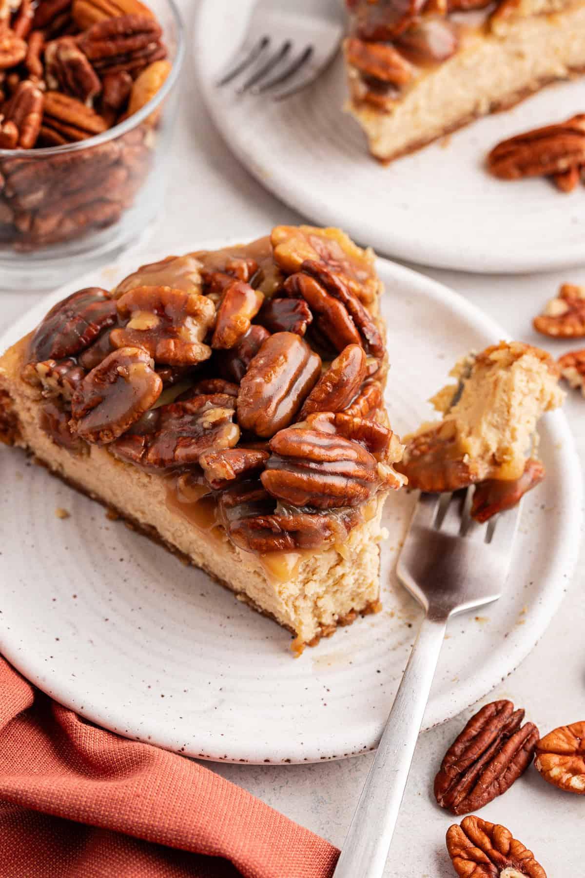 Una fetta di cheesecake alle noci pecan su un piattino bianco con una forchetta accanto e un boccone di cheesecake su di essa, circondata da un panno arancione bruciato, noci pecan sparse, una ciotola di vetro di noci pecan e un'altra fetta su un piatto separato