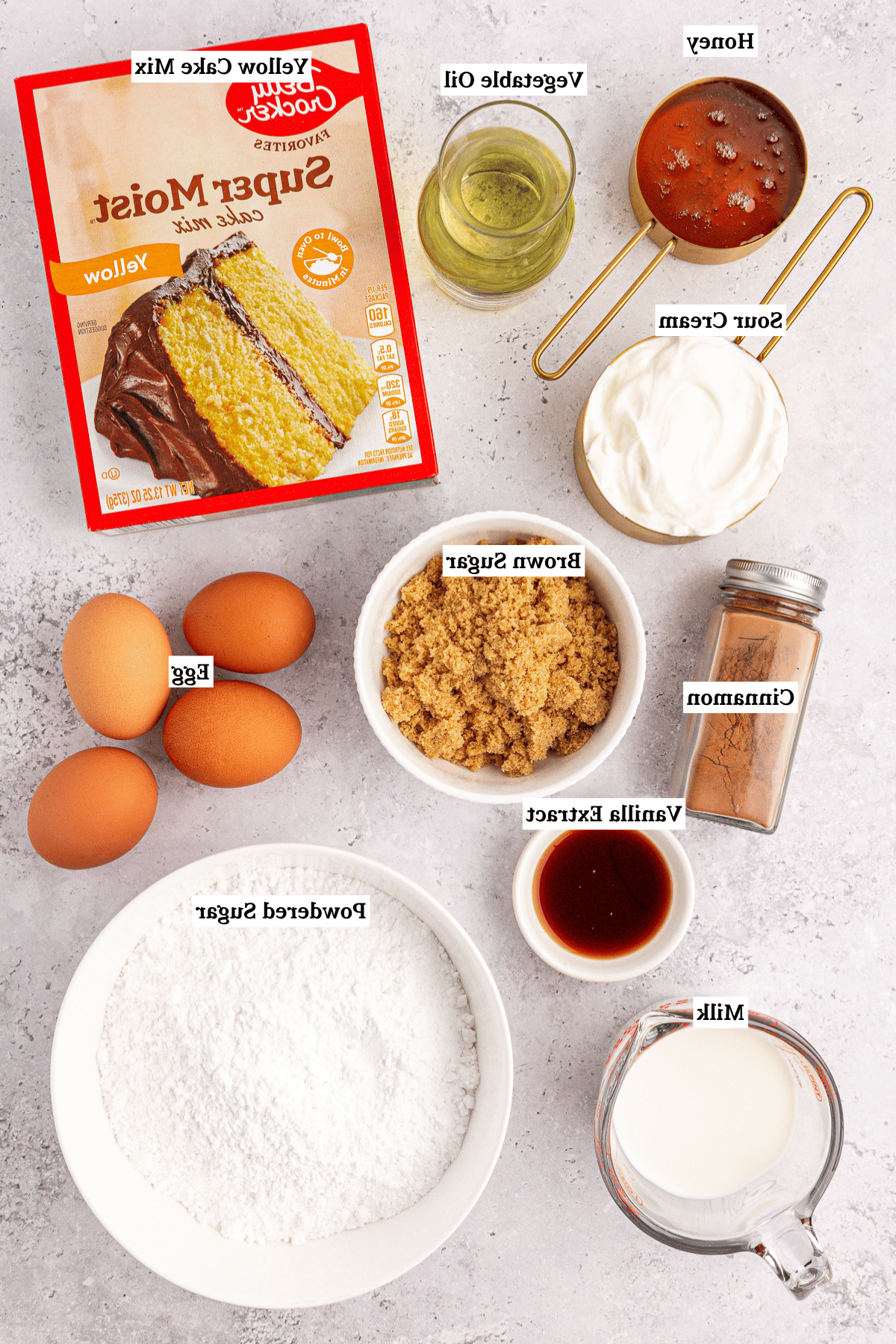 Vista dall'alto degli ingredienti per la torta Honey Bun disposti su una superficie chiara, con una scatola di preparato per torte giallo, uova, panna acida, latte, miele, olio vegetale, zucchero di canna, cannella, estratto di vaniglia e zucchero a velo.