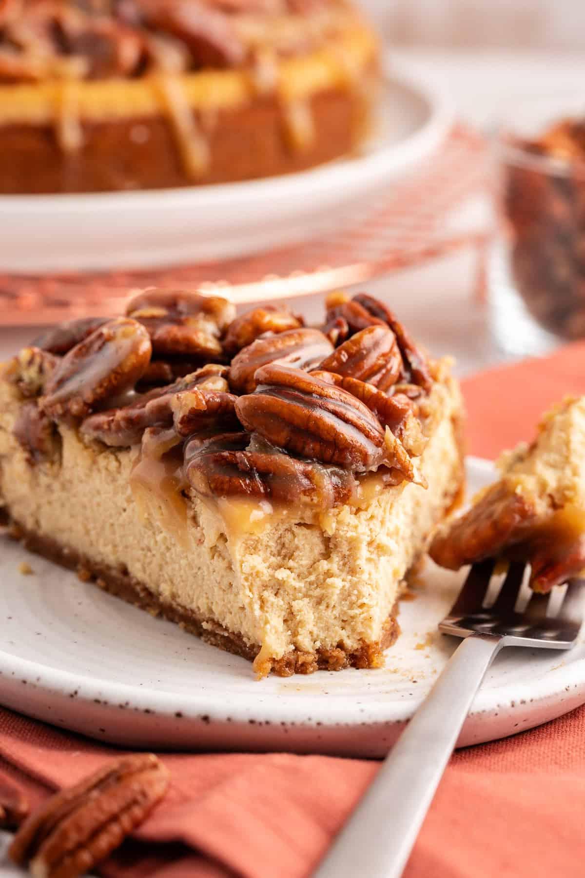Una fetta di cheesecake alle noci pecan su un piattino bianco con una forchetta con un boccone sulla parte laterale del piatto, su un panno arancione bruciato con noci pecan sparse e l'intera torta sullo sfondo