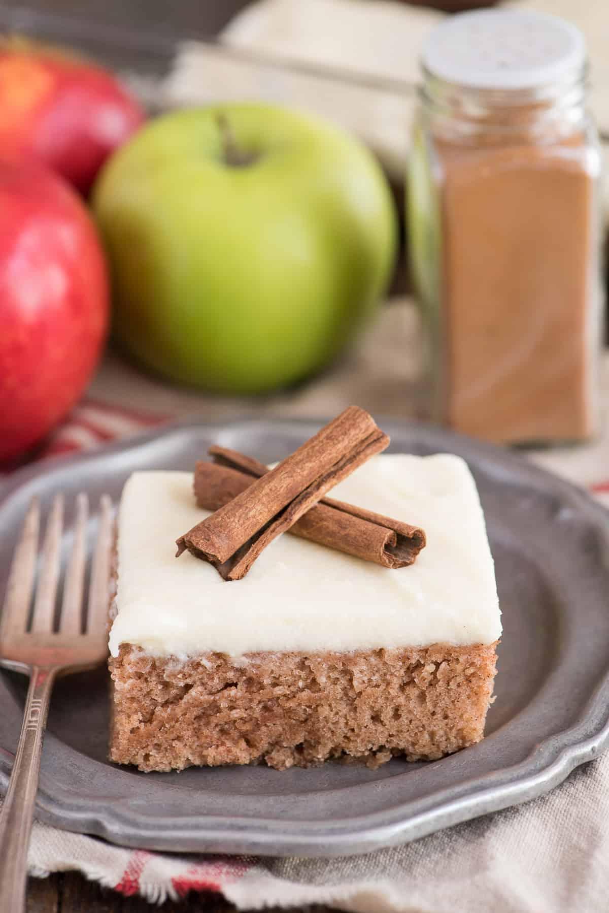 Questa torta speziata alla cannella è una ricetta autunnale incredibile! Conosciuta anche come 'wacky cake', è realizzata senza uova, latte o burro. È la torta autunnale preferita dalla nostra famiglia perché è così facile da fare! Una fetta di torta speziata alla cannella con glassa al formaggio cremoso e due stecche di cannella, su un piatto di metallo.
