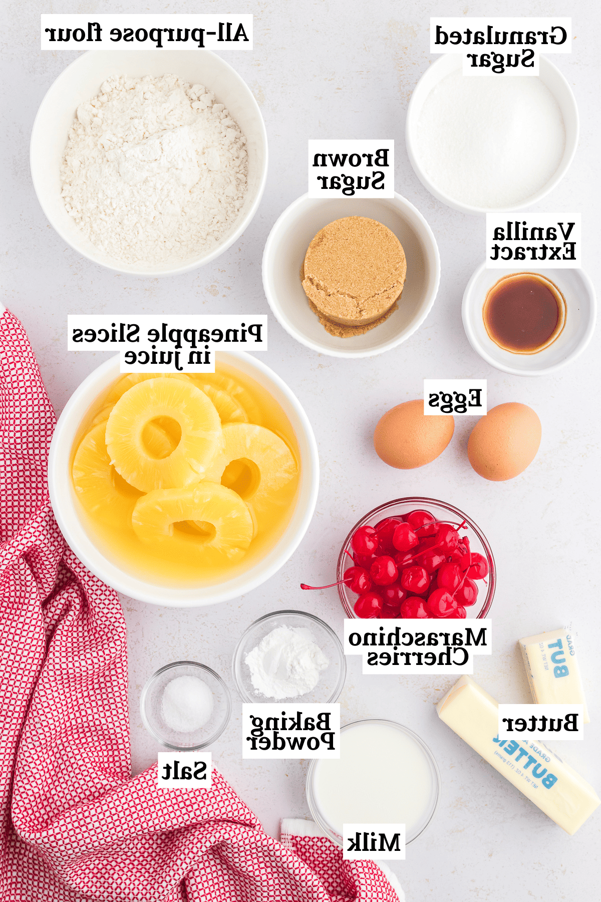 Ingredienti per la torta rovesciata all'ananas, inclusi zucchero semolato, farina tipo 00, zucchero di canna, estratto di vaniglia, uova, fette di ananas, ciliegie al maraschino, burro, lievito in polvere, sale e latte.
