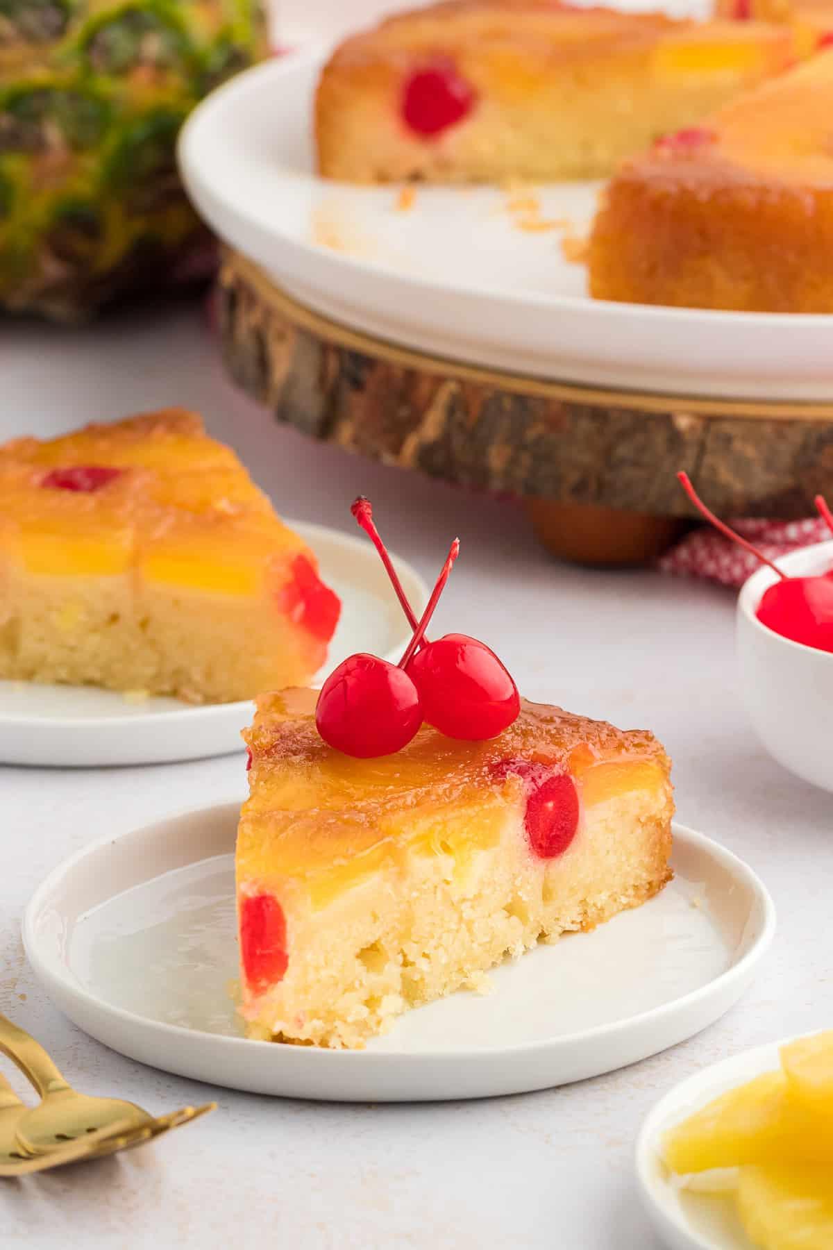 Due piccoli piatti bianchi con fette di torta rovesciata all'ananas, disposte davanti a un piatto grande con il resto della torta, due forchette dorate, fette di ananas e una piccola ciotola bianca di ciliegie.