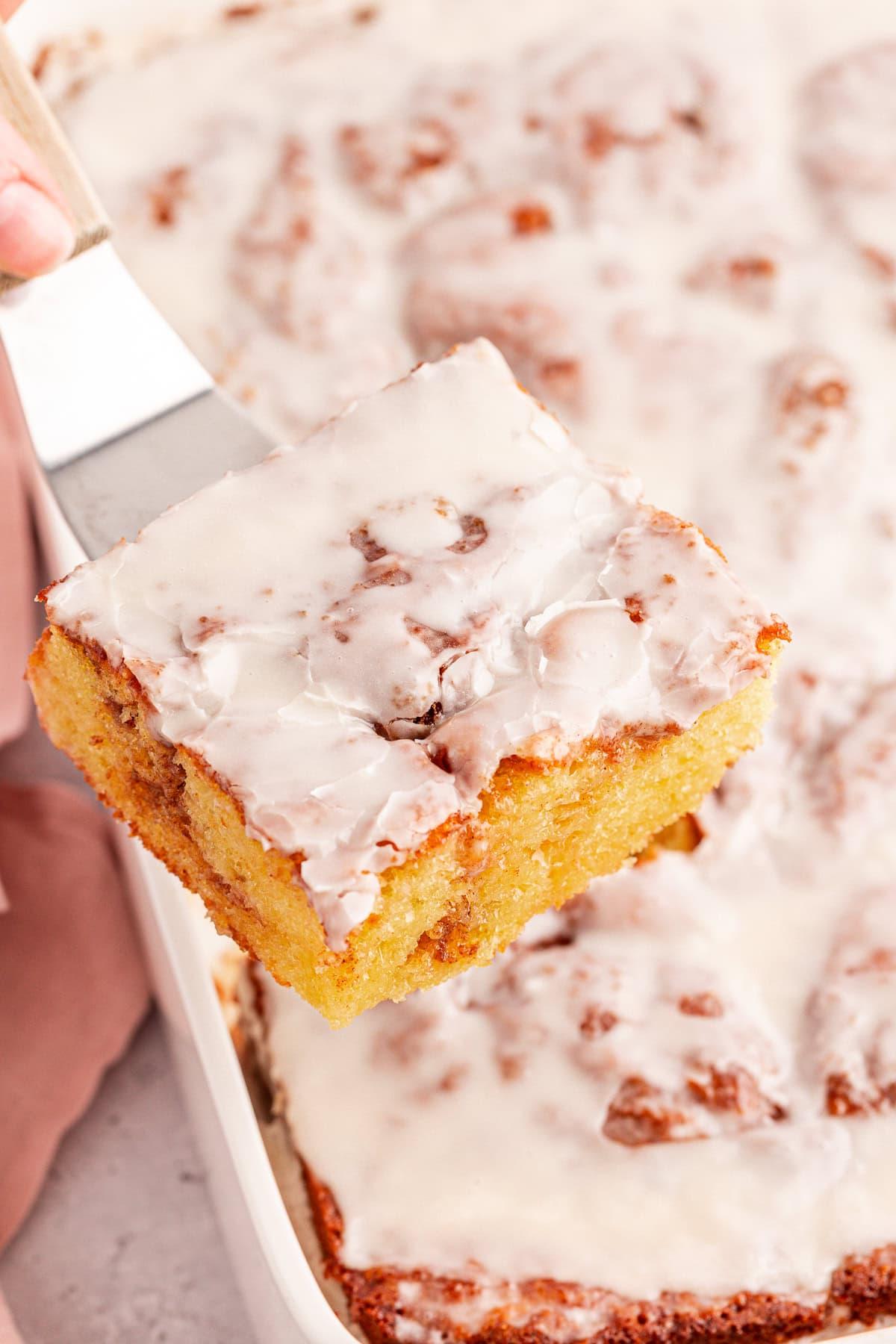 Primo piano di una fetta di torta Honey Bun glassata, sollevata da un vassoio con una spatola. La torta è dorata con una glassa bianca e visibili spirali di cannella.