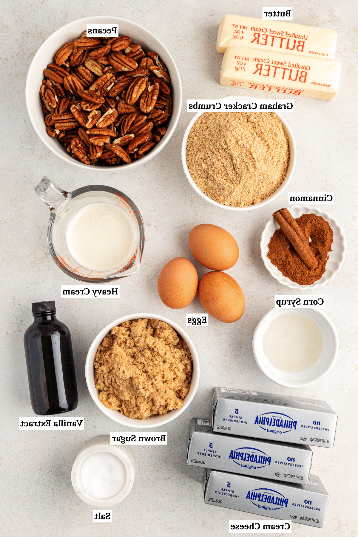Vista dall'alto degli ingredienti per la cheesecake alle noci pecan che includono burro, noci pecan, briciole di biscotti graham, cannella, sciroppo di mais, uova, panna fresca, zucchero di canna, estratto di vaniglia e sale