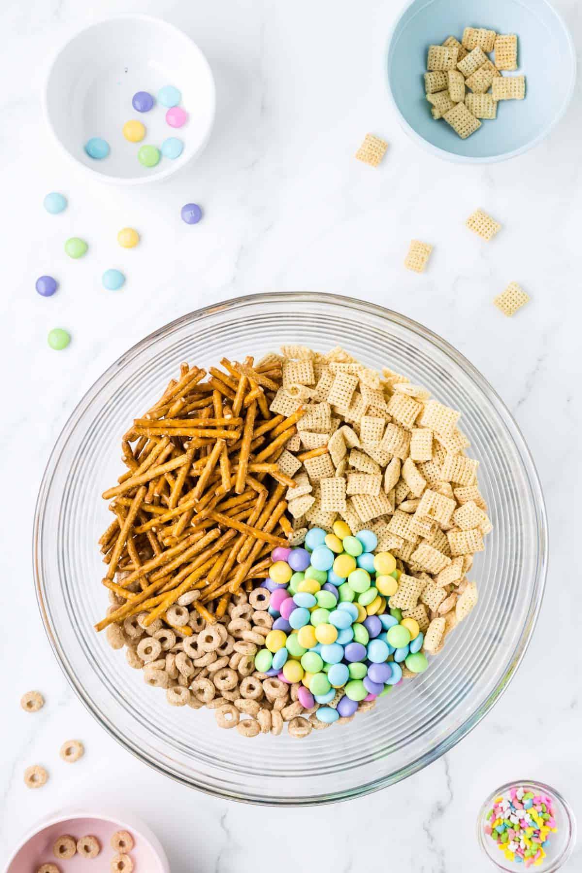 Ingredienti secchi per 'Bunny Bait' in una ciotola di vetro trasparente circondata da una ciotola di codette, una ciotola con Cheerios, una ciotola di Chex di riso e una ciotola di M&M's