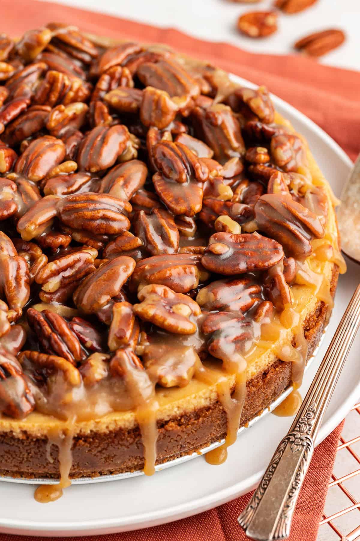 Primo piano di una cheesecake alle noci pecan su un piatto da portata bianco sopra un panno arancione bruciato su una griglia con noci pecan sparse intorno