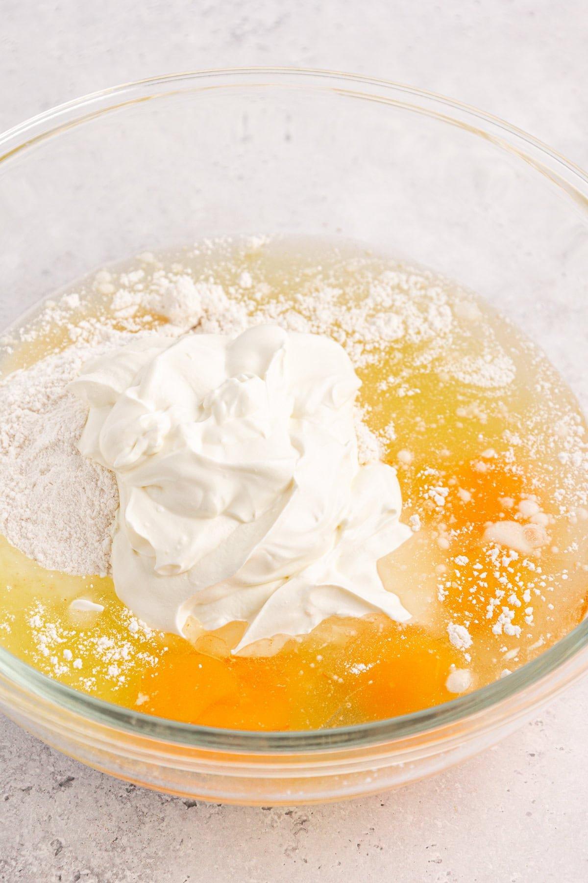 Una ciotola di vetro con ingredienti per l'impasto della torta Honey Bun contiene due uova rotte, farina, una cucchiaiata di panna acida e un accenno di cannella, pronti per essere mescolati. Lo sfondo è una superficie testurizzata grigio chiaro.