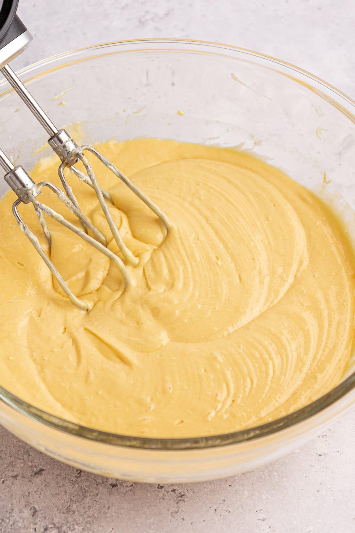 Una ciotola di vetro piena di impasto giallo liscio e cremoso, mescolato da un frullino elettrico a mano con due fruste. La superficie dell'impasto della torta Honey Bun è texturizzata con spirali dovute al processo di miscelazione. Lo sfondo è una superficie chiara e maculata.