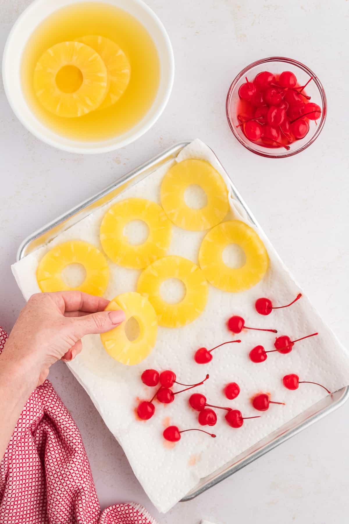 Una teglia rivestita di carta assorbente con fette di ananas e ciliegie al maraschino, accanto a un canovaccio rosso e bianco, con una mano che aggiunge un'altra fetta di ananas, accanto a una ciotola di ciliegie e una ciotola di ananas.